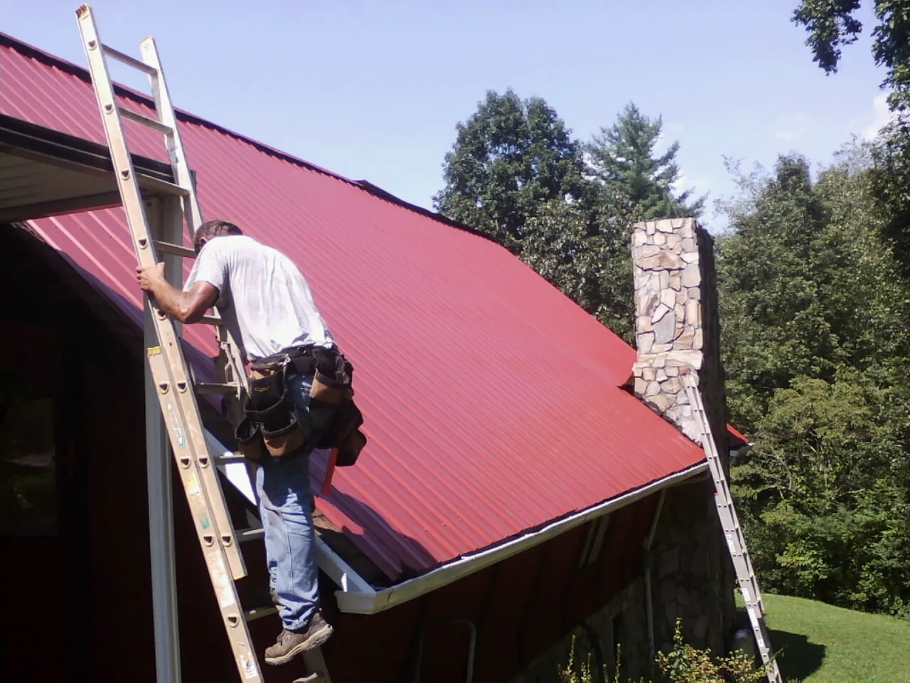 Roofer on a ladder beside a red metal roof and stone chimney — Soffit & Fascia Repair by Roofing Brothers in Island Park